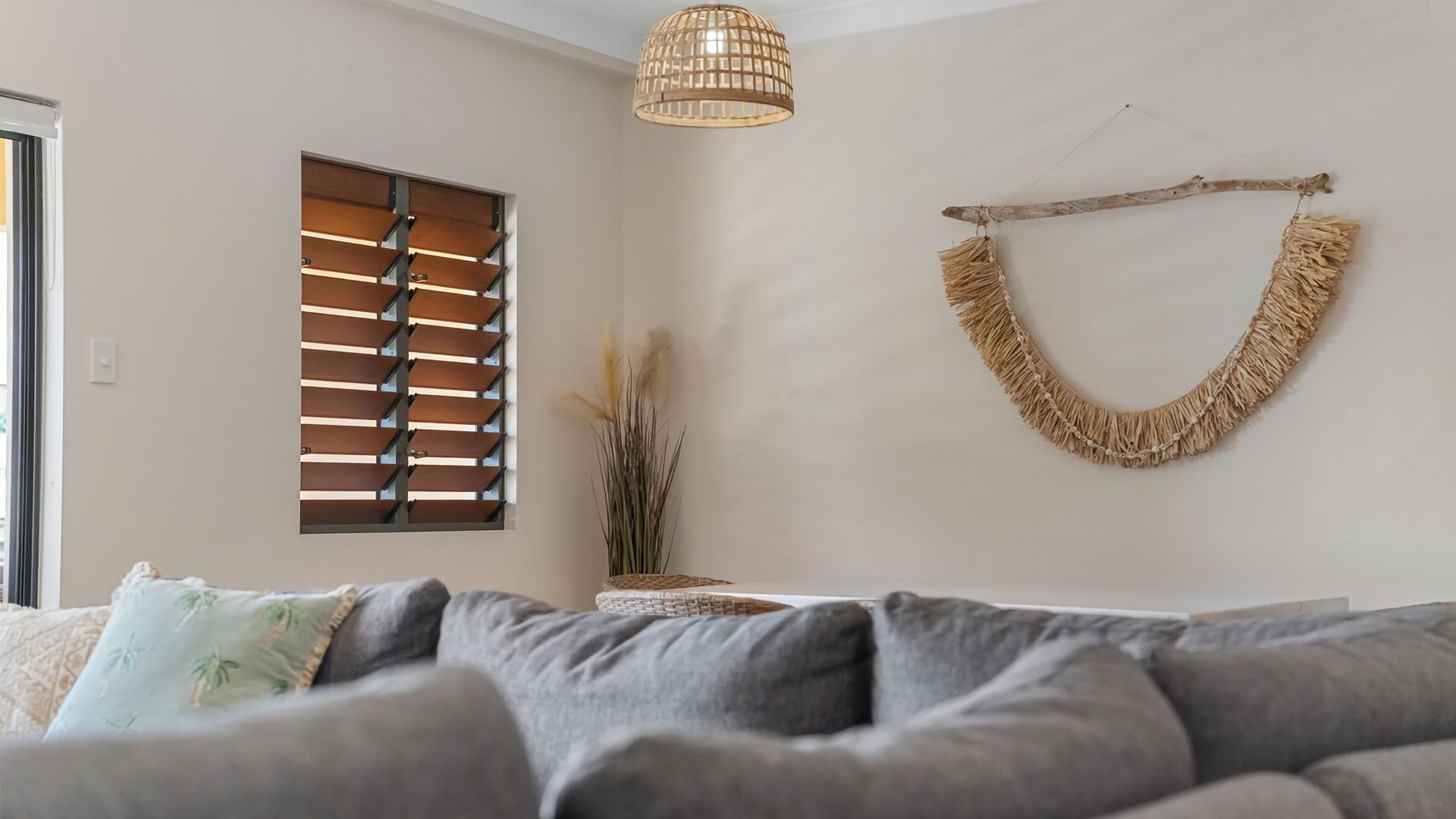A living room with a grey sofa, a window with wooden blinds, a hanging macrame wall decoration, and a pendant light.