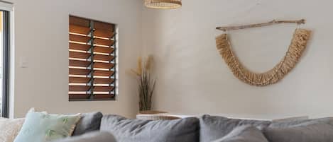 A living room with a grey sofa, a window with wooden blinds, a hanging macrame wall decoration, and a pendant light.