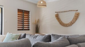 A living room with a grey sofa, a window with wooden blinds, a hanging macrame wall decoration, and a pendant light. - Seaside Stays North Ward (North Ward)