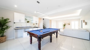 A modern living room with a pool table, a white sofa, and a kitchen area with white cabinets and a glass backsplash.