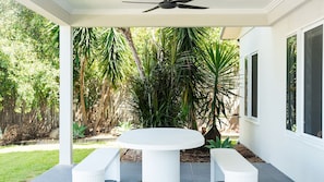 A covered outdoor area with a round table and benches, surrounded by lush greenery and a wooden fence.