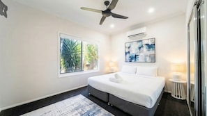 A modern bedroom with a bed, a ceiling fan, an air conditioning unit, a window with a view of greenery, and a painting on the wall.