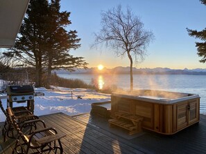 Eine Holzterrasse mit Whirlpool und Grill, mit Blick auf einen See, im Hintergrund Berge.