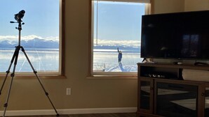 A room with large windows overlooking a snowy landscape, a tripod with a camera, and a television on a wooden stand. - Shorebird Cottage (Homer)