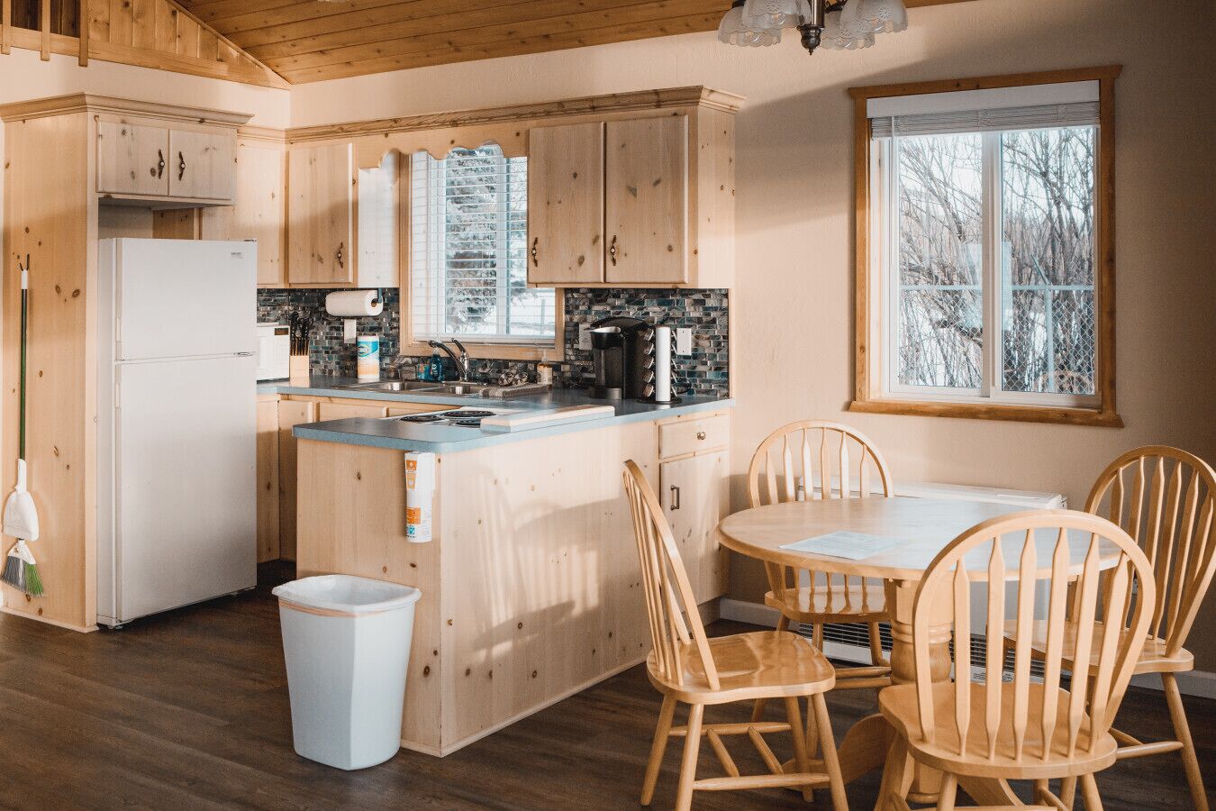 A kitchen with wooden cabinets, a refrigerator, a sink, and a window with a view of trees.