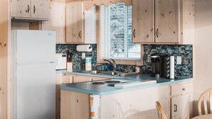A modern kitchen with wooden cabinets, a white refrigerator, a sink, and a window with a view of trees. - Shorebird Cottage (Homer)