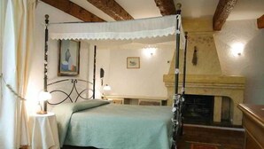 A four-poster bed with a green bedspread, a small table with a lamp, a painting on the wall, and a fireplace. - Le Logis du Guetteur (Les Arcs-sur-Argens)