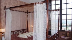 A four-poster bed with a canopy, a bedside table with a lamp, and a view of the outdoors through a window. - Le Logis du Guetteur (Les Arcs-sur-Argens)