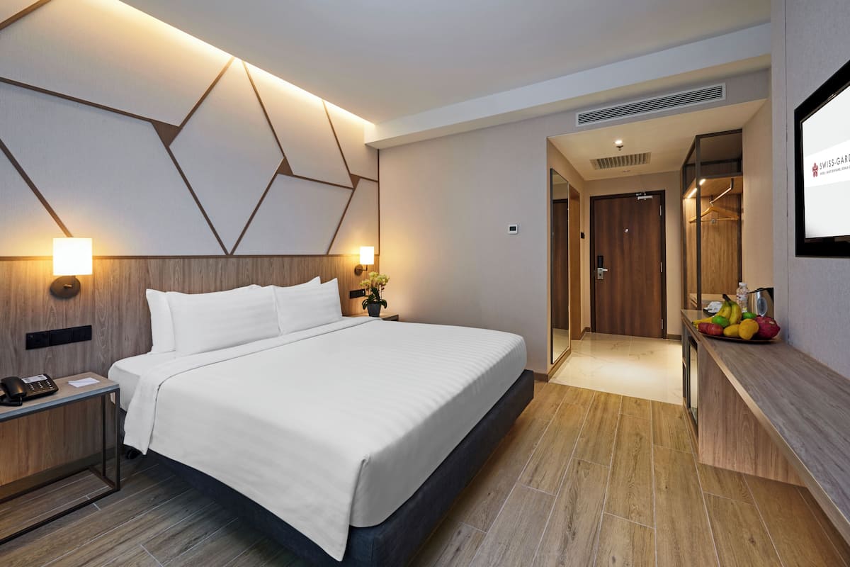 Ein modernes Hotelzimmer mit einem großen Bett, einem Nachttisch mit Telefon, einem Beistelltischchen mit Lampe, einer Obstschale und einem Spiegel.