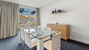 A hotel room with a glass table and chairs, a wooden cabinet, and a view of a body of water and hills. - Family-Friendly 3BR Marina Views & Space (Queenstown)