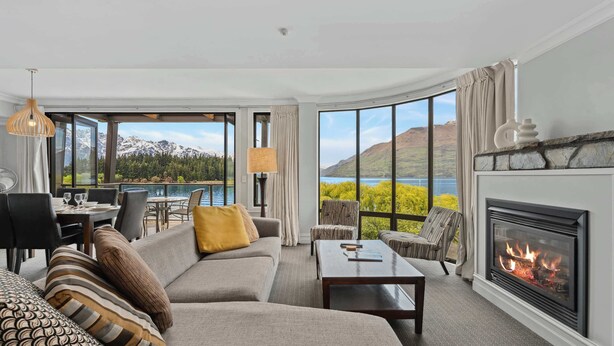 A modern living room with a fireplace, large windows with a mountain view, and a dining area. - Gorgeous Lakefront, 3 Bed apartment, walk to town (Queenstown)
