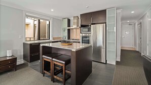 A modern kitchen with dark cabinetry, a central island with stools, stainless steel appliances, and a large window. - Gorgeous Lakefront, 3 Bed apartment, walk to town (Queenstown)