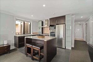 A modern kitchen with dark cabinetry, a central island with stools, stainless steel appliances, and a large window. - Gorgeous Lakefront, 3 Bed apartment, walk to town (Queenstown)