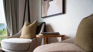 Ein modernes Wohnzimmer mit einer Couch, einem Sessel mit Kissen, einem kleinen Holztisch mit Büchern und einem gerahmten Kunstwerk an der Wand.