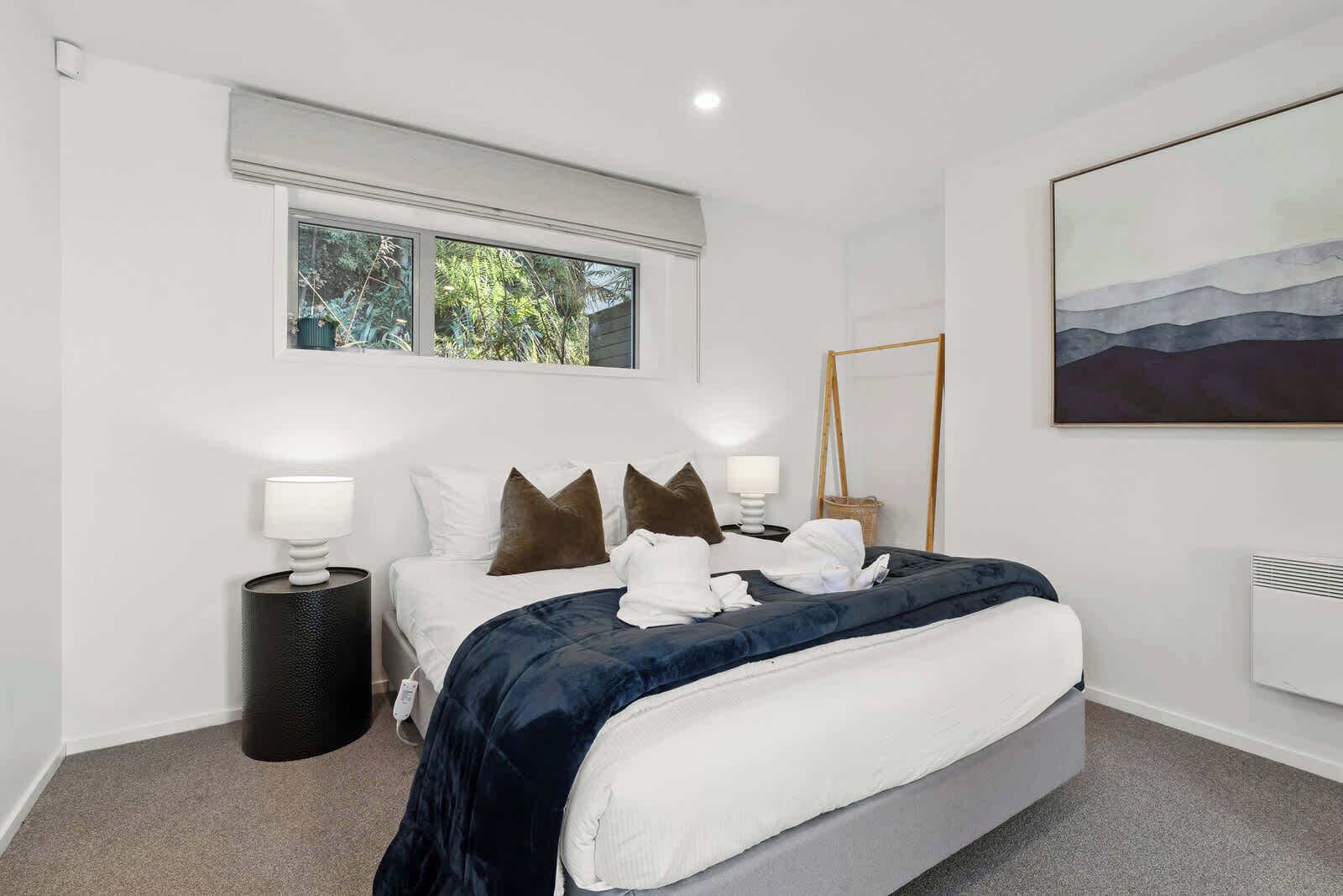 A modern bedroom with a large bed, two bedside tables, a window, and a painting on the wall.