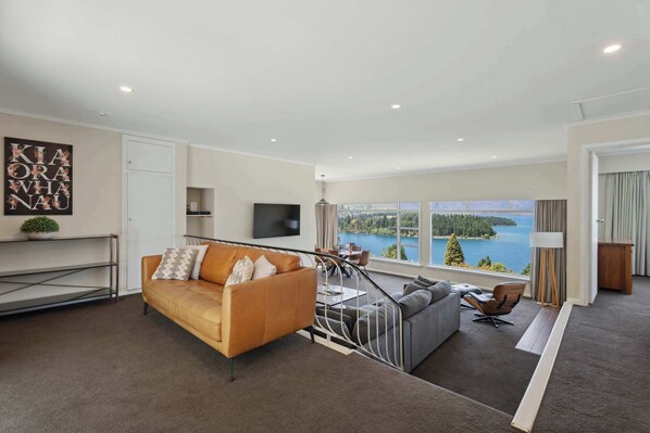 A spacious living room with a sofa, a grey couch, and a staircase leading to another level. A TV is mounted on the wall, and there is a large window offering a view of a lake and mountains. - The Biggest & Best Views, 3 Bedrooms, Walk To Town (Queenstown)