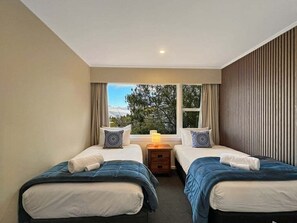 A hotel room with two beds, a window with curtains, a bedside table with a lamp, and a view of trees outside. - The Biggest & Best Views, 3 Bedrooms, Walk To Town (Queenstown)
