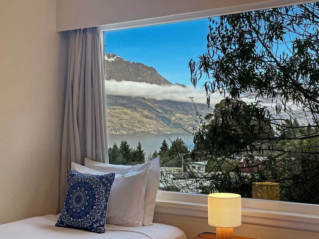 A bedroom with a bed, a window with curtains, and a view of mountains and a lake.