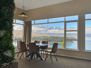 A dining area with a table and chairs, large windows with a mountain view, and a hanging light fixture. - The Biggest & Best Views, 3 Bedrooms, Walk To Town (Queenstown)
