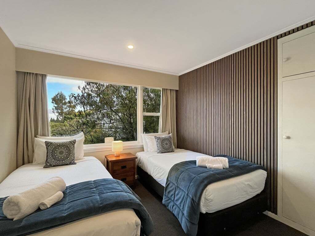 A hotel room with two beds, a window with curtains, a bedside table with a lamp, and a wardrobe.
