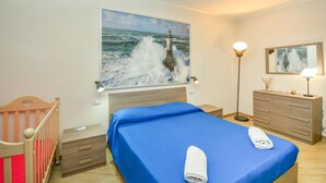 Una camera da letto con un quadro grande raffigurante un faro, un letto con biancheria azzurra, un comò in legno e un comodino con una lampada.