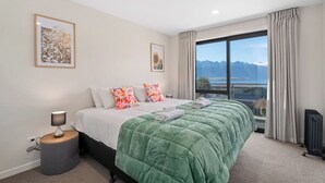 A bedroom with a large bed, a bedside table with a lamp, a view of mountains through a window, and two framed pictures on the wall. - Quiet Home with Stunning Lake & Mountain Views (Queenstown)