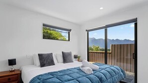 A bedroom with a large bed, a nightstand, a window with a view of mountains, and a sliding glass door to an outdoor area. - Central Charm | 10 Min Walk to CBD (Queenstown)