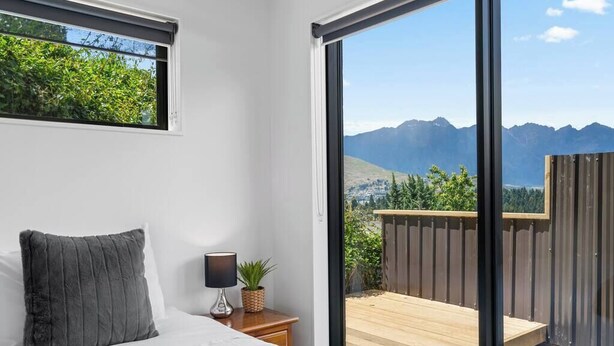 A bedroom with a bed, a nightstand, a lamp, and a view of mountains through a sliding glass door. - Central Charm | 10 Min Walk to CBD (Queenstown)
