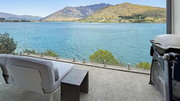 Un balcon avec vue sur un lac et des montagnes, un canapé gris et un barbecue.