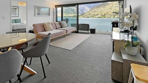 Un salon moderne doté d’une grande fenêtre donnant sur un lac et des montagnes, avec un canapé, une table à manger et des chaises, ainsi qu’un petit bureau agrémenté d’une plante.