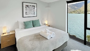 Une chambre avec un grand lit, une table de chevet, une commode, un tableau encadré au mur et une vue sur un lac et des montagnes, visible par une grande fenêtre.