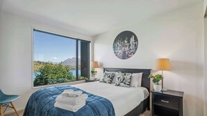 Un dormitorio con una cama grande, vista a una montaña y un lago, un cuadro redondo y una mesita de noche con una lámpara.