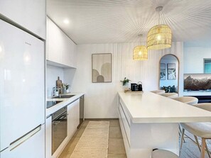 A modern kitchen with white cabinetry, a built-in oven, a large island with seating, and pendant lighting. - Charming Retreat | Balcony, Views and Garage (Queenstown)