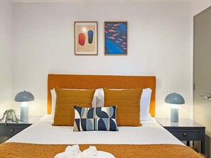 Ein Hotelzimmer mit einem großen Bett, zwei Nachttischen mit Lampen und zwei gerahmten Kunstwerken an der Wand.