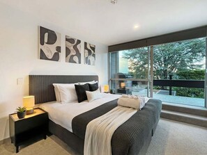Ein modernes Schlafzimmer mit einem großen Bett, einem Nachttisch, einer Lampe und Blick ins Freie durch ein großes Fenster.