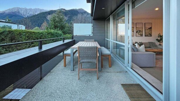 Un balcón moderno con mesa y sillas, una puerta de cristal que da a un interior con un sofá y obras de arte, y una vista de la montaña al fondo.
