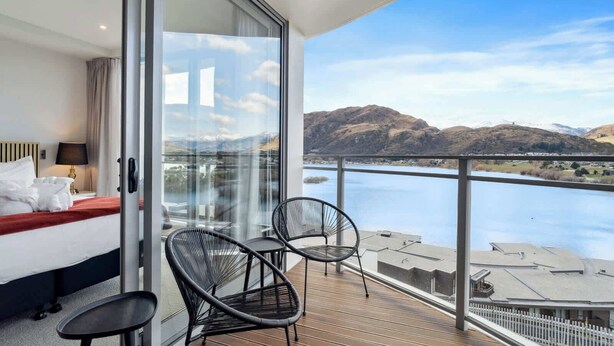 A hotel room with a bed, a glass door leading to a balcony with chairs and a view of a lake and mountains. - Sunlit Serenity, Lakefront Apartment (Kawarau Falls)
