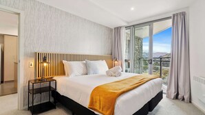 A modern hotel room with a large bed, a nightstand with a lamp, and a view of the mountains through the window. - Sunlit Serenity, Lakefront Apartment (Kawarau Falls)