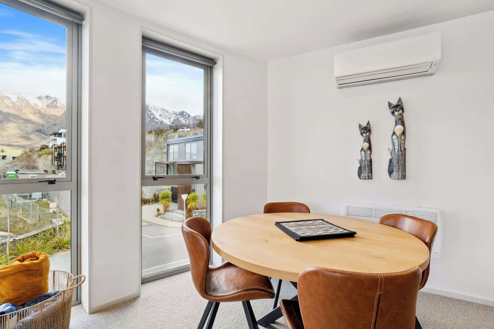 A modern dining area with a round table and leather chairs, a large window with a mountain view, and two wall-mounted cat sculptures.