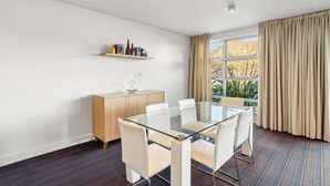 A modern hotel room with a glass dining table, white chairs, a wooden cabinet, and a large window with curtains. - Waterfront 3 Bedroom Marina Gem (Queenstown)
