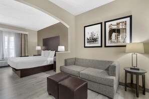 Una camera d'albergo con un letto, un divano grigio, un poggiapiedi marrone e due quadri appesi al muro.