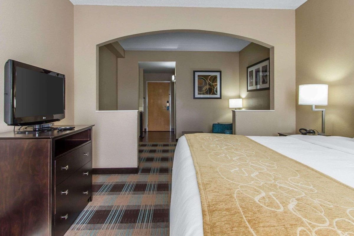 A hotel room with a bed, a television on a dresser, a lamp, and a door leading to a hallway.