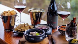 Une table dressée avec divers aliments et boissons, notamment des verres à vin, une bouteille de vin et des bols remplis de nourriture, le tout placé devant une fenêtre donnant sur des arbres.