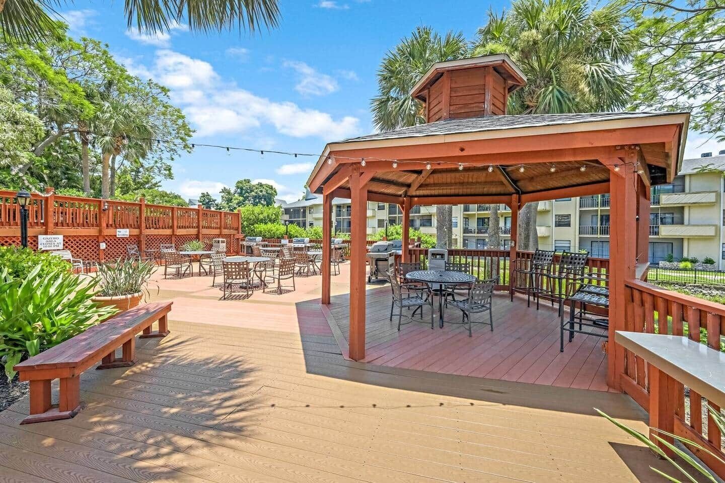 Un'area patio esterna con tavoli e sedie in legno, una zona relax coperta e vista su un edificio residenziale sullo sfondo.
