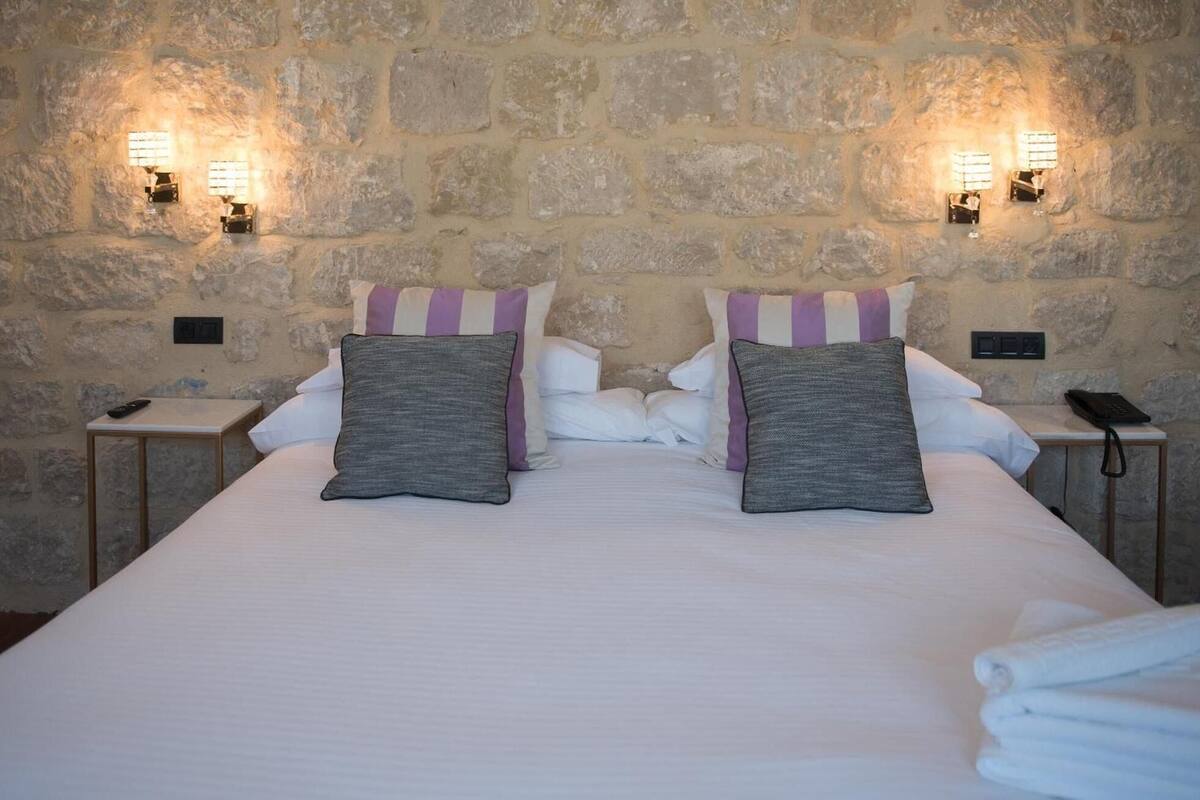Una habitación de hotel con pared de piedra, una cama con sábanas blancas, dos almohadas con fundas a rayas y mesitas de noche con lámparas y teléfonos.