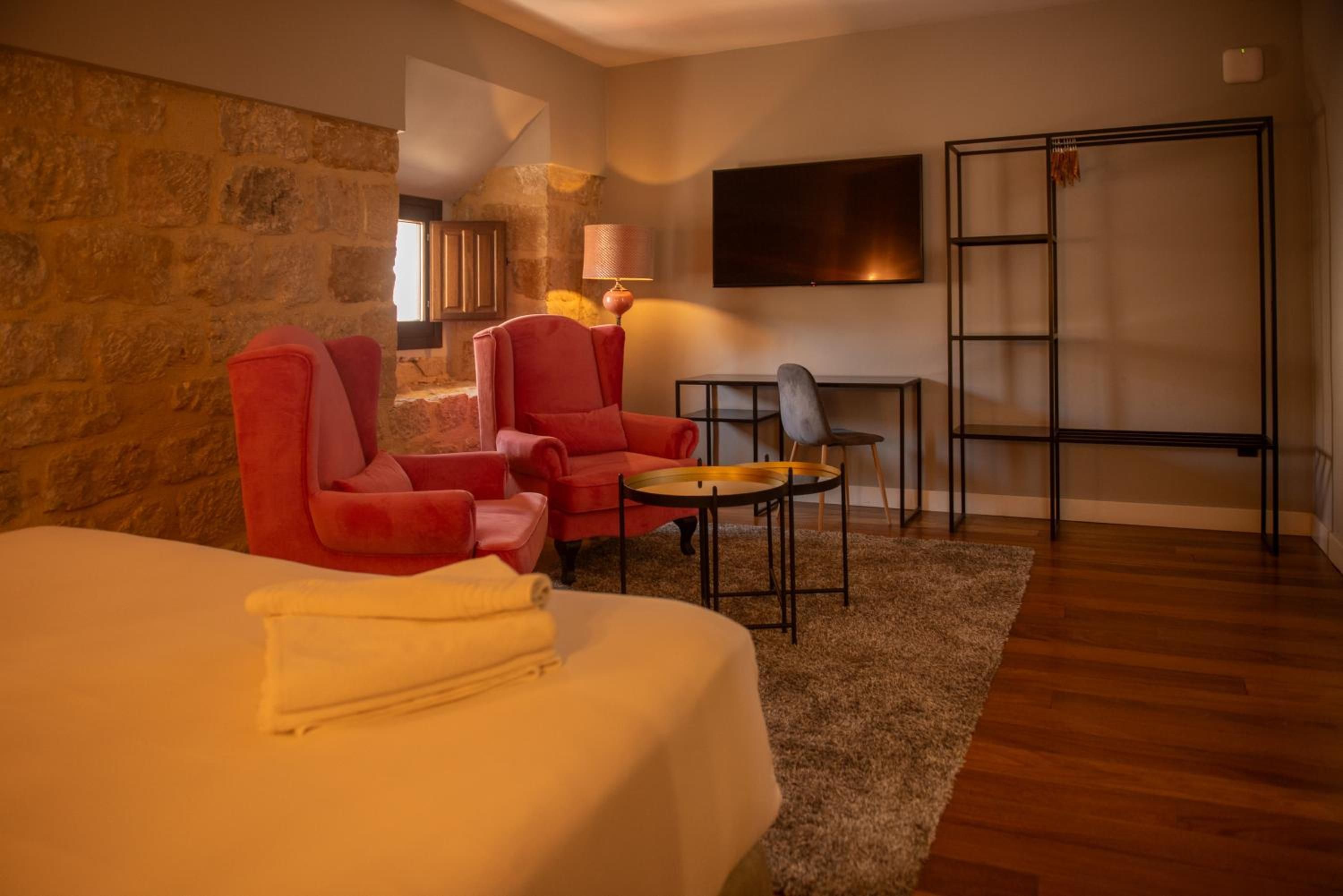 A hotel room with a bed, two red armchairs, a small table, a TV, and a wooden floor.