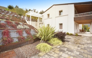 Une maison moderne dotée d’un patio, d’un espace extérieur aménagé et couvert, et d’un jardin paysager agrémenté de plantes et d’un petit élément décoratif aquatique.