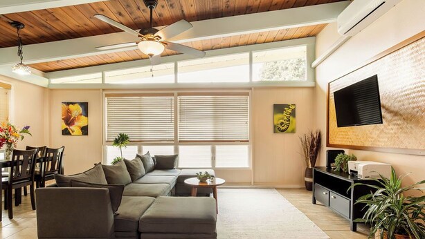 A modern living room with a sectional sofa, a ceiling fan, a flat-screen TV mounted on the wall, and a dining area with chairs and a table. - Hale Hibiscus (Hilo)