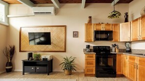 A modern kitchen with wooden cabinets, a black stove, and a microwave. There is a large flat-screen TV mounted on the wall, a small table with a plant, and a hanging light fixture. - Hale Hibiscus (Hilo)