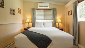 A hotel room with a large bed, two bedside tables with lamps, a window with blinds, and an air conditioning unit. - Hale Hibiscus (Hilo)